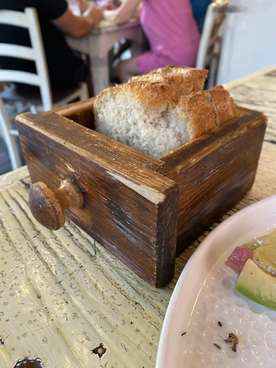 A small wooden drawer holds slices of bread on a rustic table. A plate with avocado and other food is partially visible, and people sit in the blurry background.