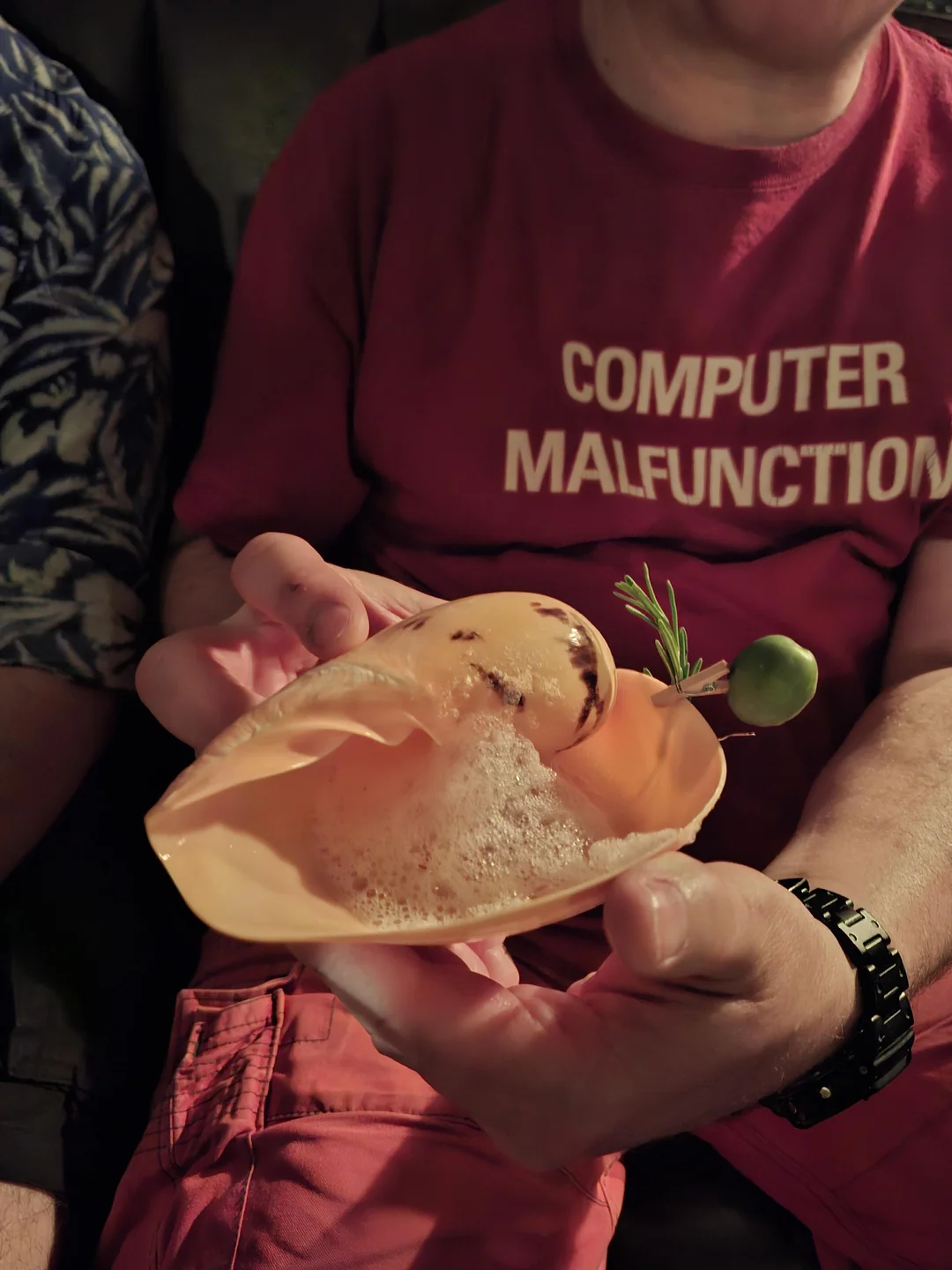 A person in a red “COMPUTER MALFUNCTION” shirt holds a shell-shaped dish with foam, a grilled garnish, a green olive, and a sprig of rosemary.