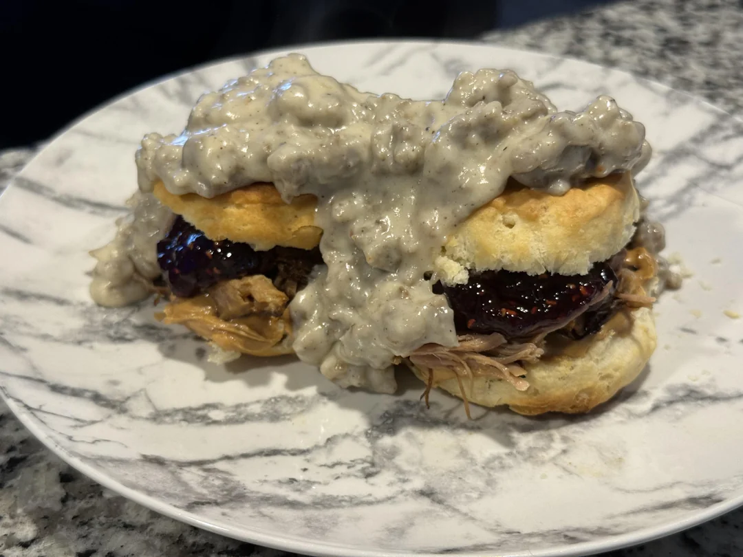 A biscuit sandwich filled with pulled pork and barbecue sauce, topped with sausage gravy, served on a white plate with a gray marble pattern.