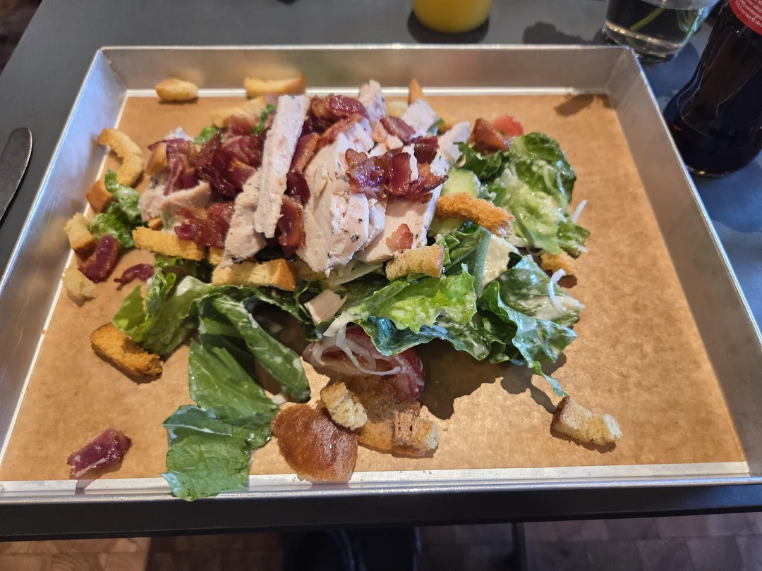 A Caesar salad with grilled chicken strips, romaine lettuce, croutons, grated cheese, and bacon bits, served on a metal tray lined with brown paper. Drinks are visible in the background.