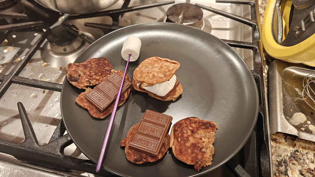 A plate on a stove holds three mini pancake s’mores, each with chocolate and marshmallow. A skewer with a toasted marshmallow rests on the plate. Nearby is a yellow oven mitt and a pan.