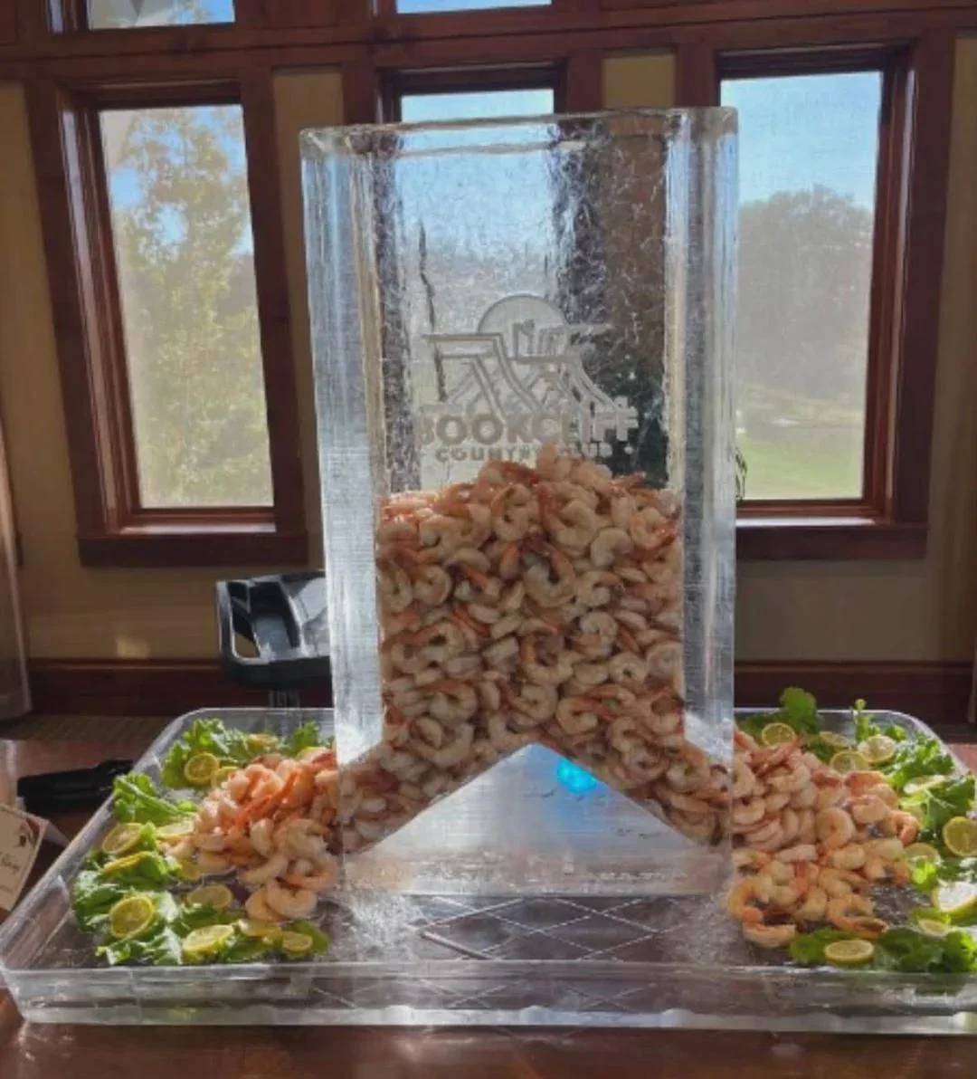 Shrimp piled in and around a tall, clear ice sculpture with an engraved logo. Lettuce and lemon slices decorate the base. Sunlight streams through windows in the background.