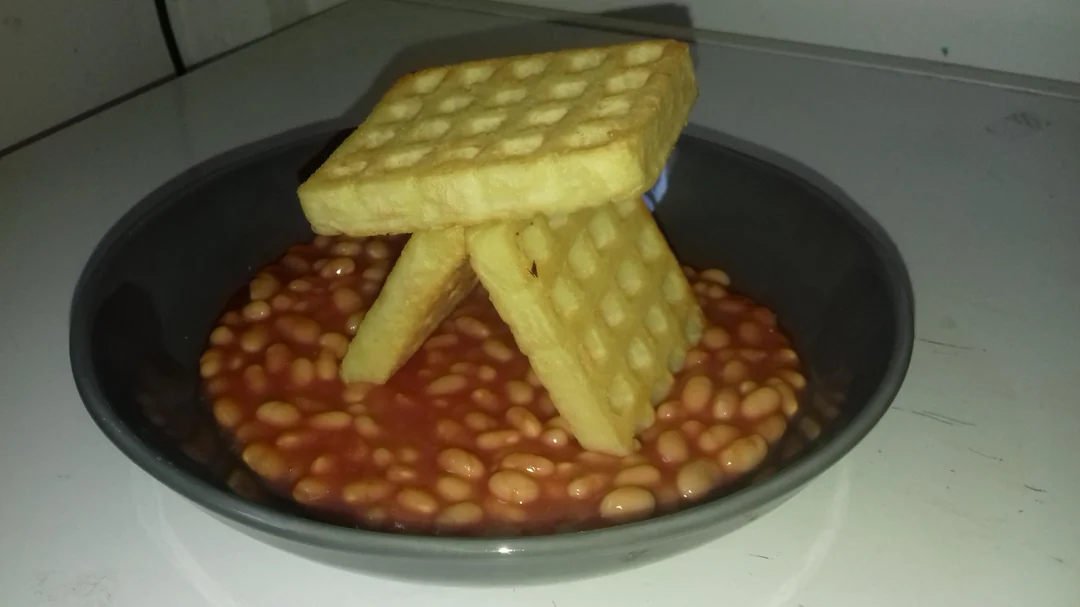Unusual food photos. A black bowl of baked beans with two potato waffles arranged in a tent shape on top. The bowl sits on a white surface.