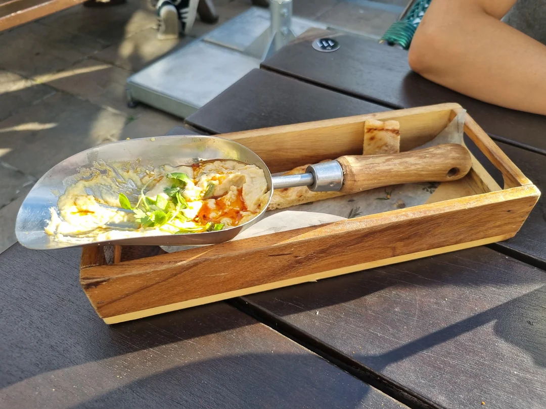 A metal scoop with leftover dip and herbs rests in a wooden box, with a piece of flatbread beside it, on a dark outdoor table in the sunlight.