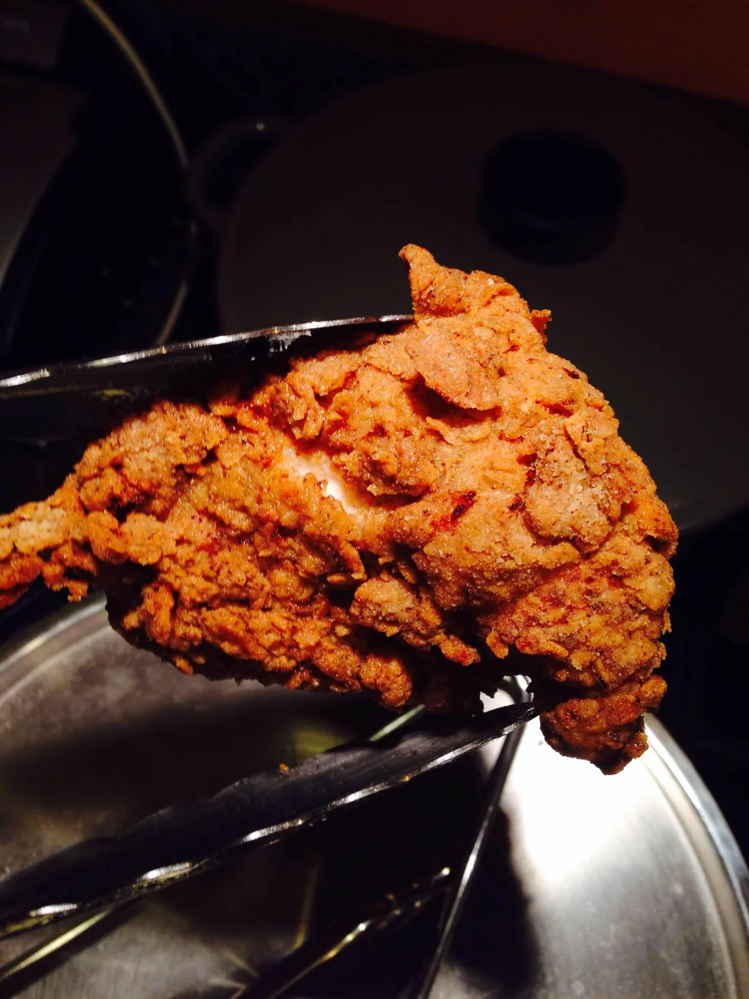 A piece of crispy fried chicken held by metal tongs above a reflective metal surface, with warm lighting highlighting its golden brown texture.