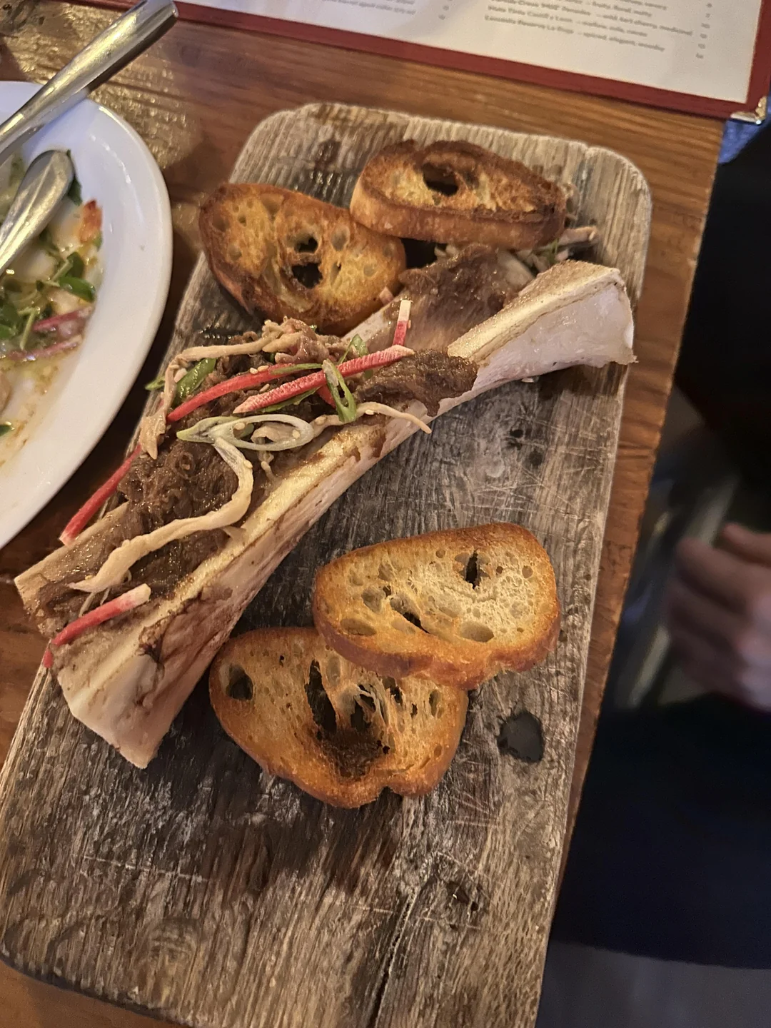 Roasted bone marrow topped with thinly sliced vegetables, served on a rustic wooden board with toasted bread slices on the side.