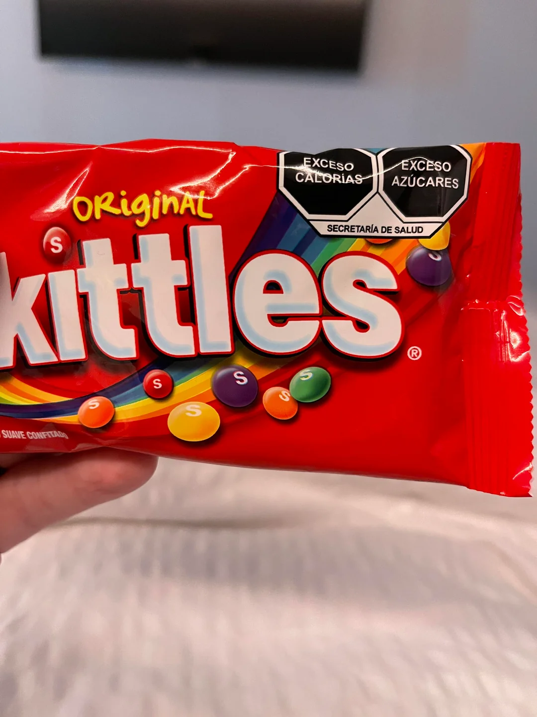 A close-up of a red Skittles candy package with Spanish labels warning “Exceso Calorías” and “Exceso Azúcares.” Multiple colorful Skittles are displayed on the packaging.