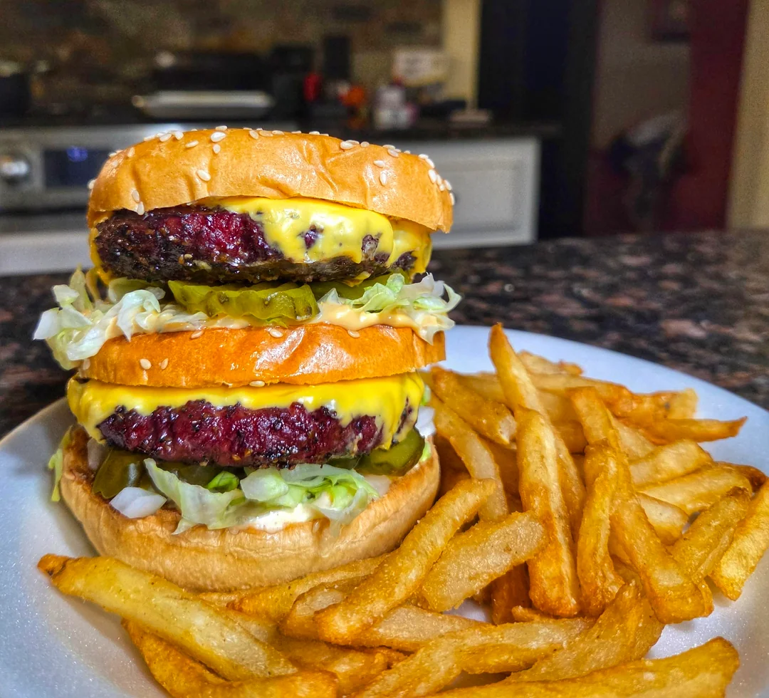 A double cheeseburger with lettuce, pickles, and two beef patties, served on a white plate with a side of golden French fries. The background shows a kitchen setting.