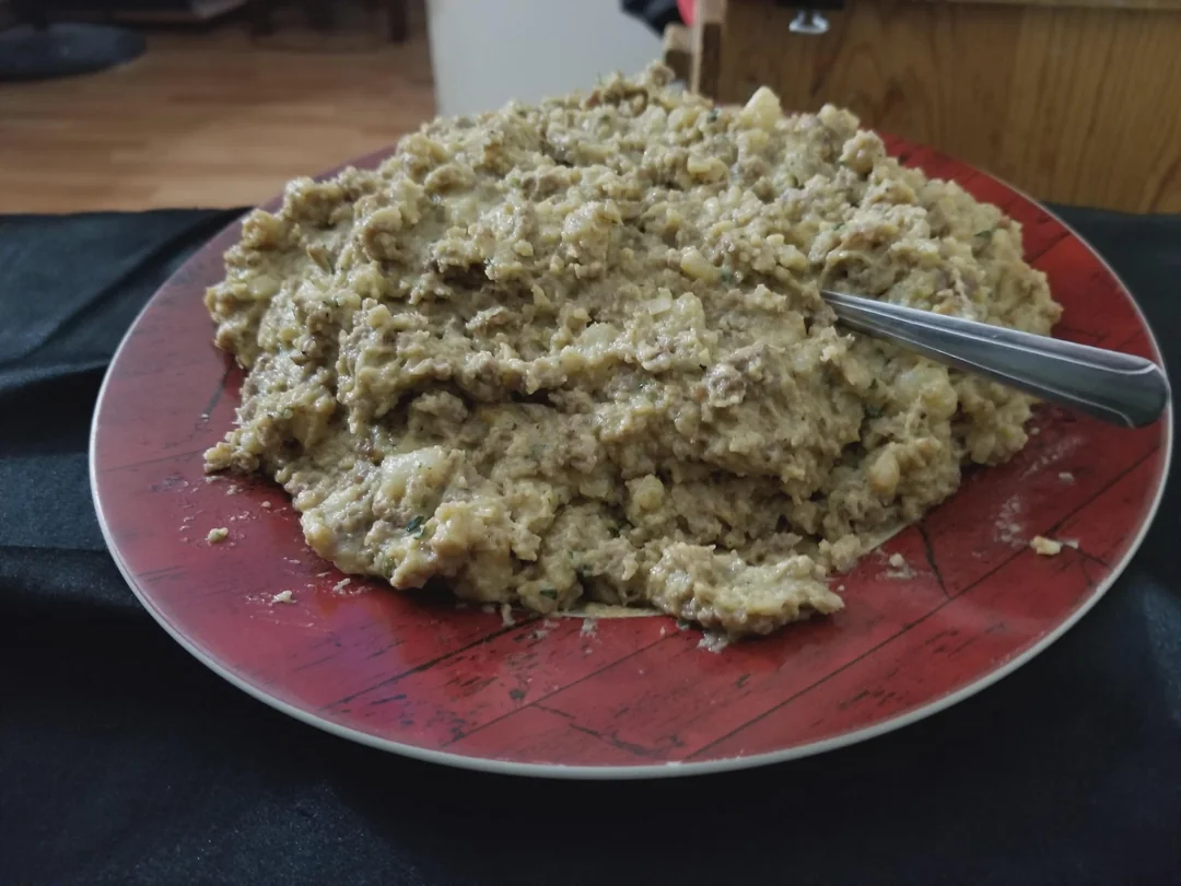 A red plate piled with a large serving of lumpy, beige-colored food, possibly mashed potatoes or stuffing, sits on a black surface. A spoon is partly embedded in the food.