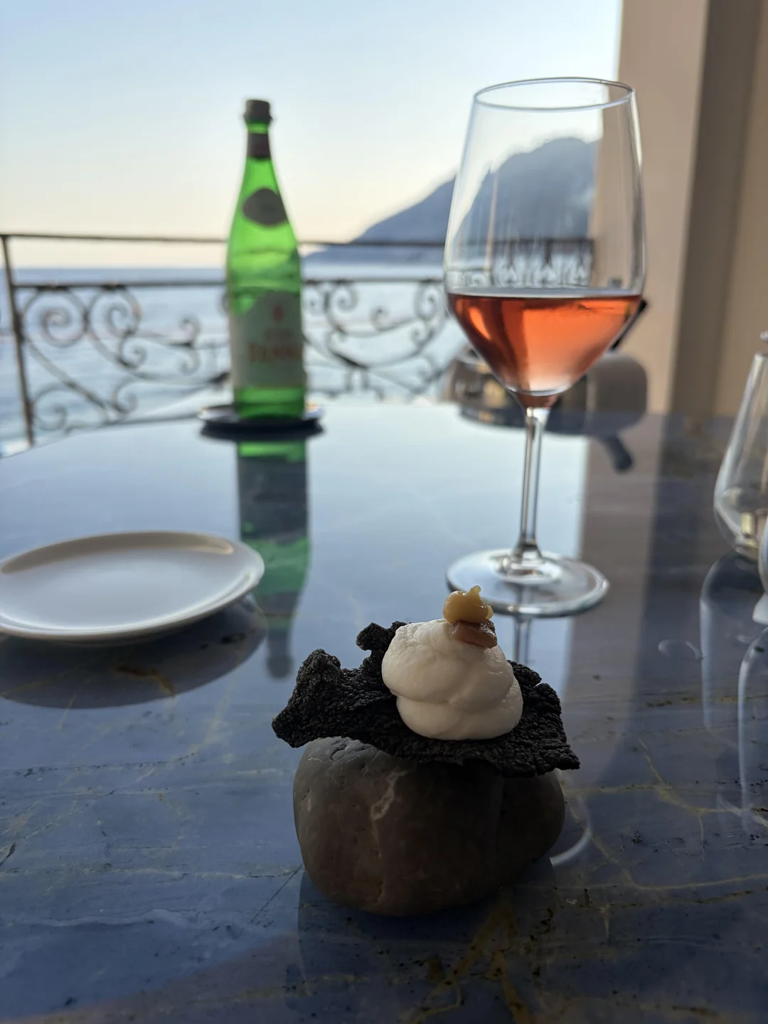A glass of rosé wine and a green bottle sit on a table overlooking the sea, with a small appetizer on a rock in the foreground. A mountain and ornate railing are visible in the background.
