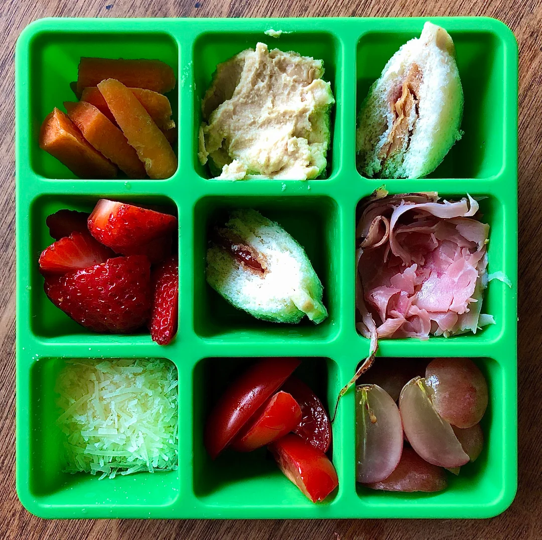 A green tray with nine compartments, each holding different foods: sliced plums, hummus, sandwich pieces, strawberries, prosciutto, shredded cheese, tomatoes, and grapes on a wooden table.