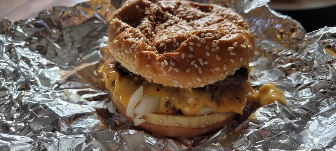 A cheeseburger with melted cheese, onions, and a sesame seed bun sits on crumpled aluminum foil.