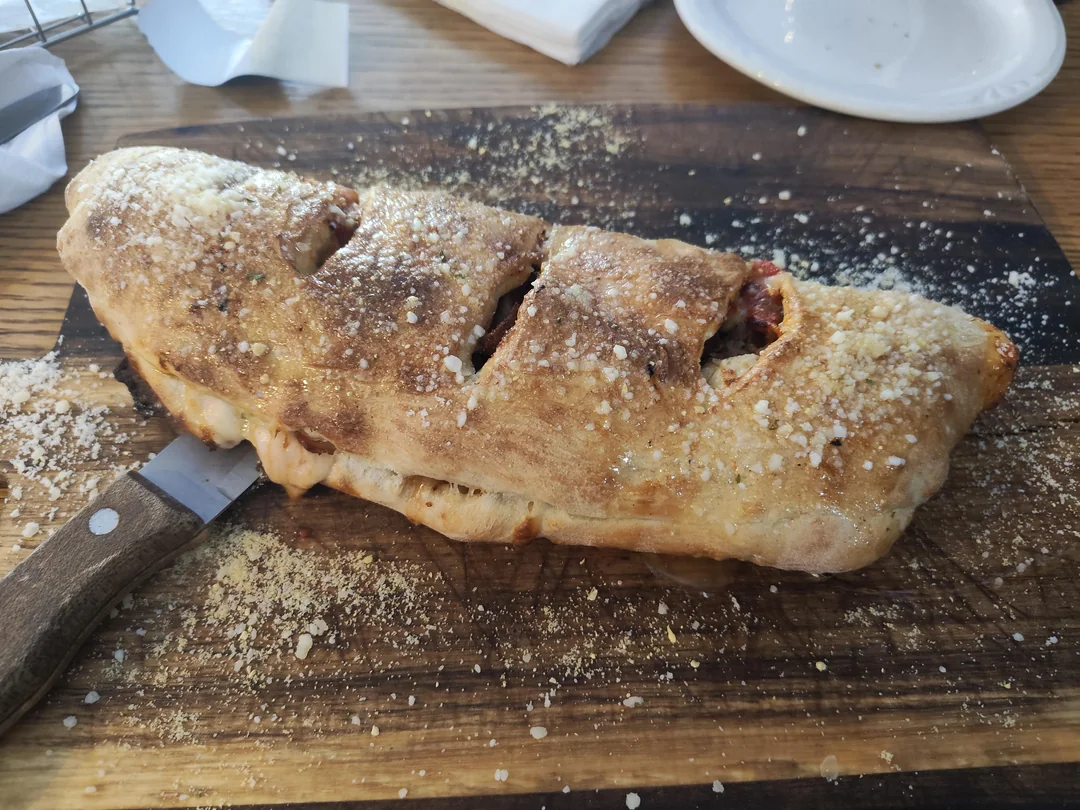 A baked calzone sprinkled with grated cheese sits on a wooden board, partially sliced open with a knife resting beside it. A white plate and napkin are in the background.