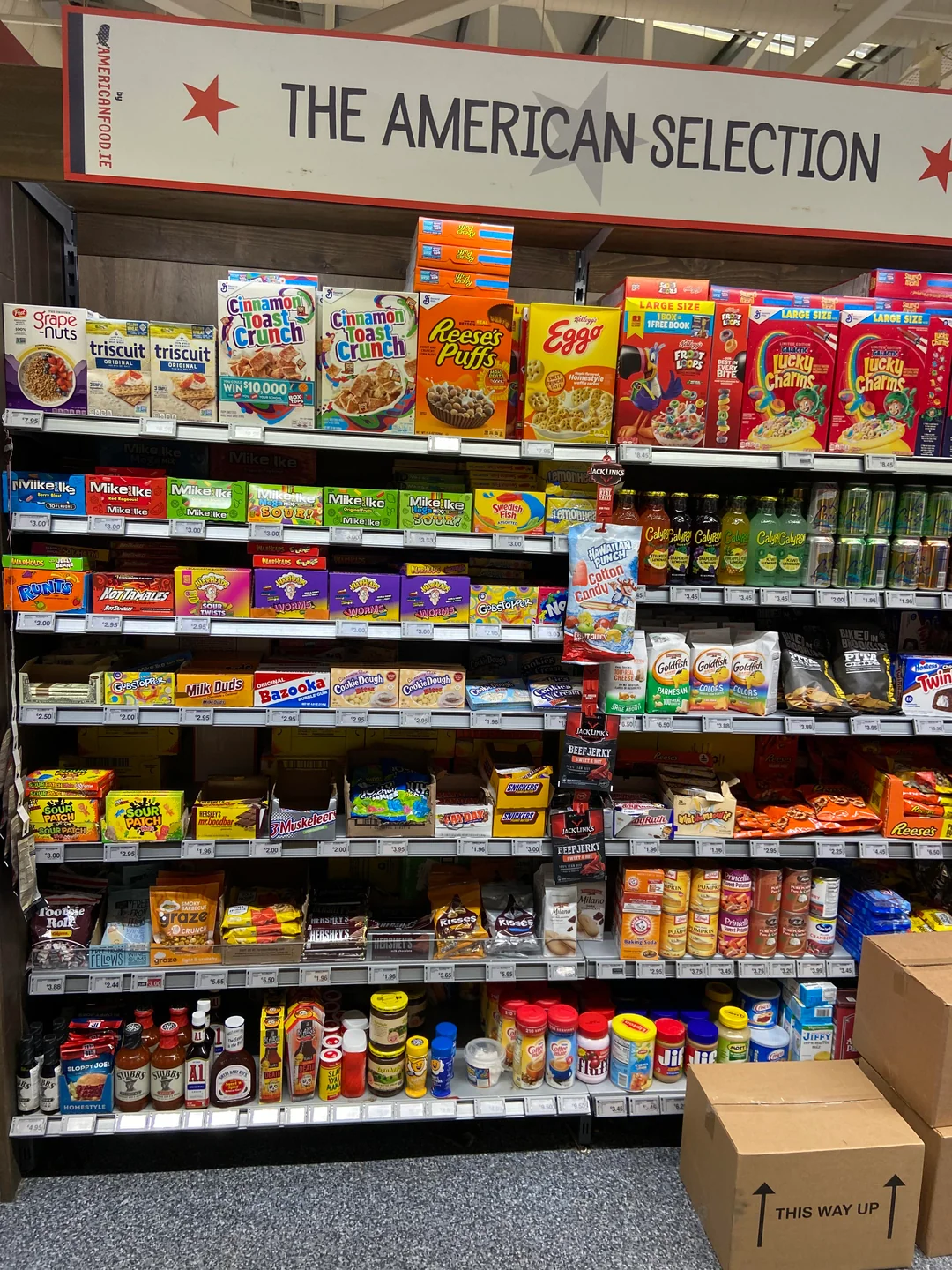 Shelves labeled “The American Selection” display various American cereals, snacks, Pop-Tarts, peanut butter, baking goods, and sauces in a grocery store. Some boxes and jars are stacked with visible brand logos.