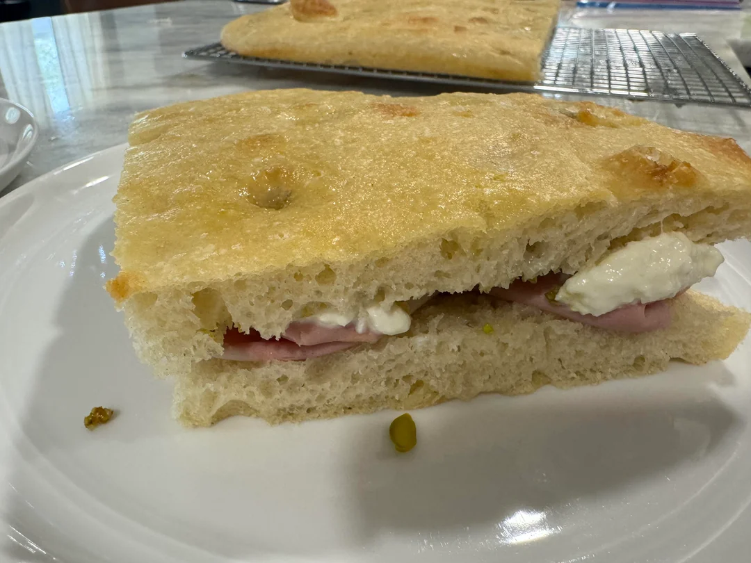 Fast food copycat meals. A sandwich made with thick focaccia bread, filled with slices of ham and mozzarella cheese, sits on a white plate. Another piece of focaccia is visible in the background on a cooling rack.