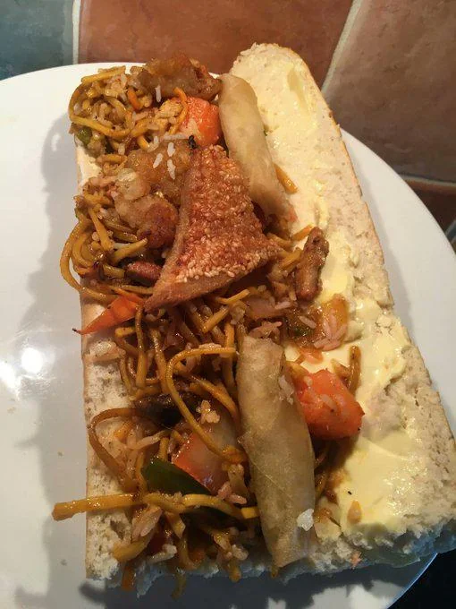 A sandwich roll filled with stir-fried noodles, rice, vegetables, spring roll pieces, and pork crackling, with butter spread inside, on a white plate.
