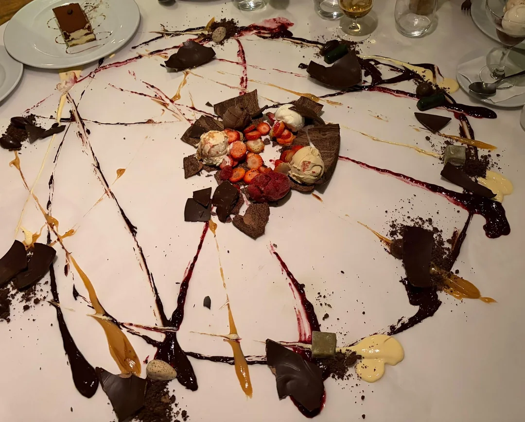 A dessert is artistically spread on a large table, featuring sauces, strawberries, ice cream, cake, and chocolate, arranged in a circular pattern on white paper.