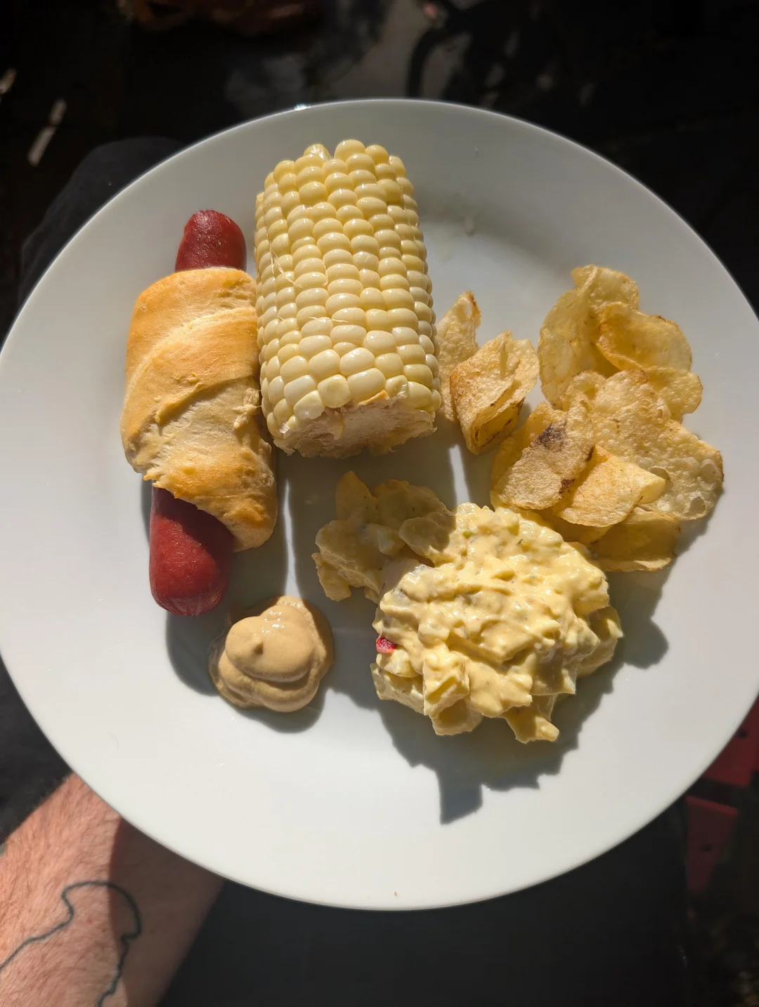 A plate with a crescent roll-wrapped hot dog, corn on the cob, a serving of potato chips, a scoop of potato salad, and a dollop of mustard.