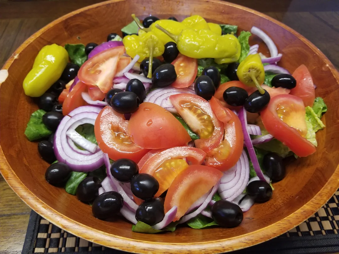 A wooden bowl filled with salad, featuring sliced tomatoes, red onions, black olives, pepperoncini peppers, and leafy greens.