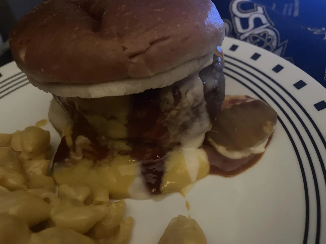 A cheeseburger with melted cheese and sauce sits on a plate next to a serving of macaroni and cheese. The burger appears juicy and slightly messy, with cheese dripping down the sides.