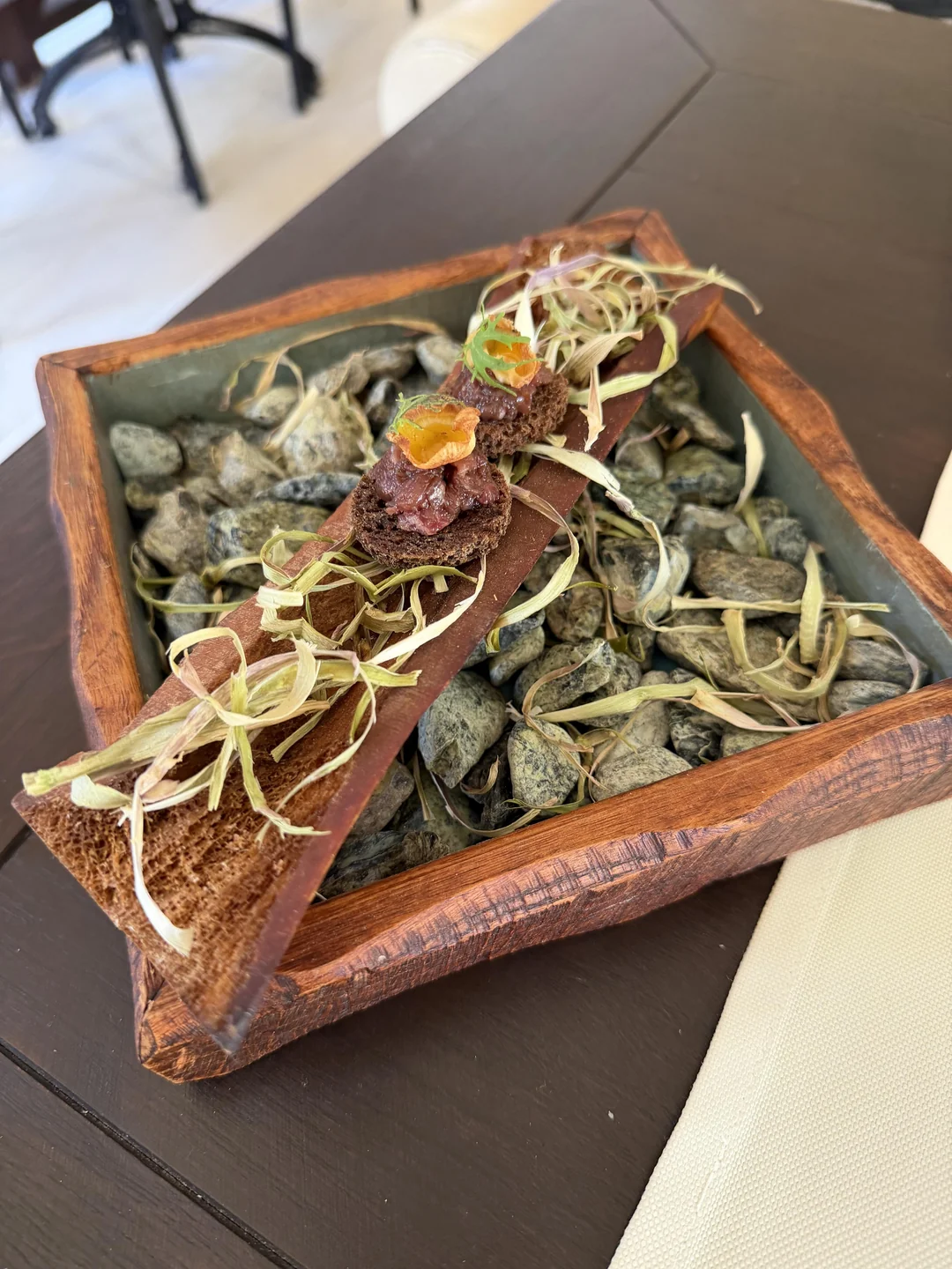 A wooden box filled with stones and straw holds a piece of bark topped with gourmet food garnished with herbs, presented as a rustic, artistic appetizer on a dark tabletop.