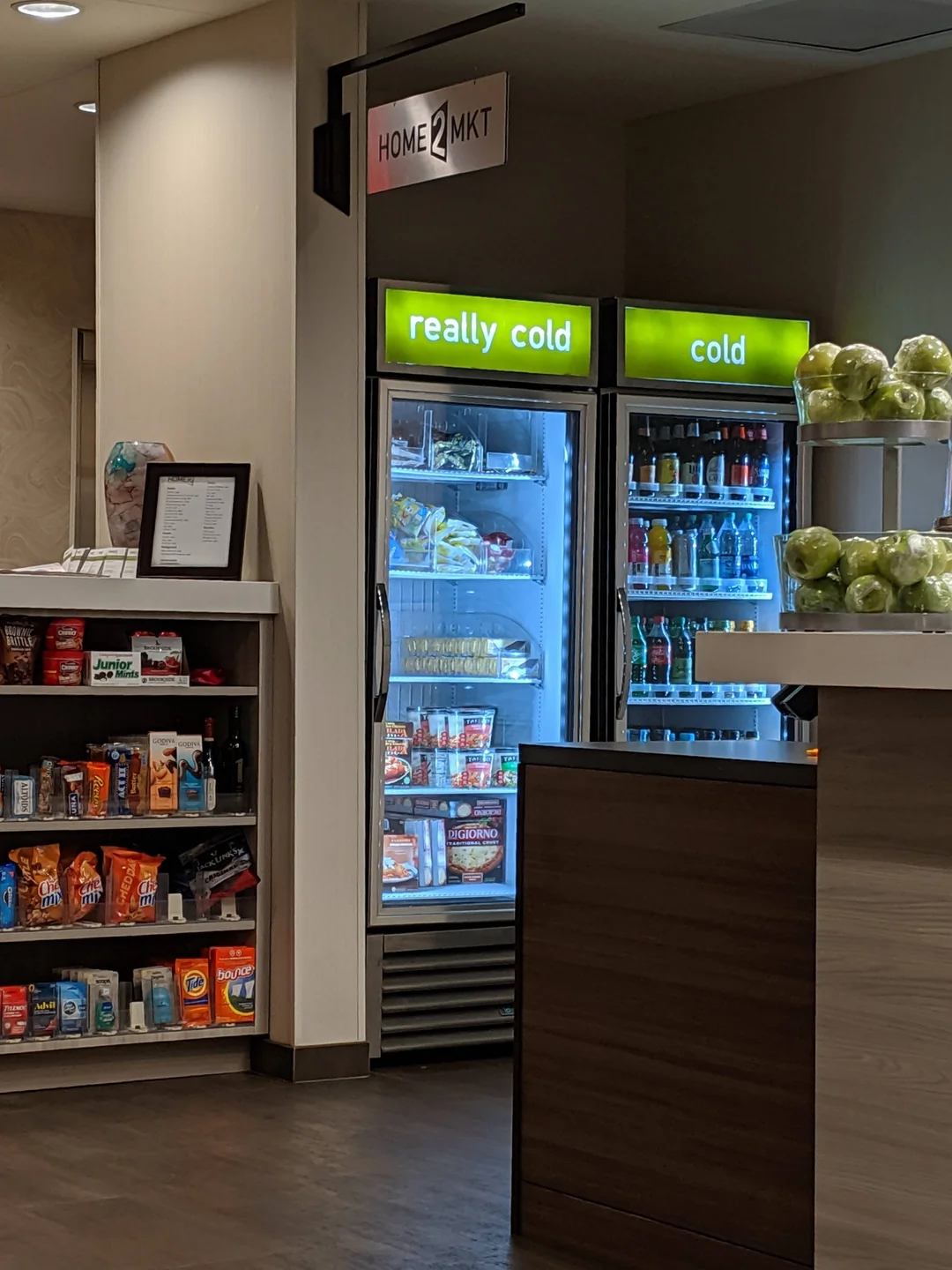 A hotel market area with a snack shelf, a counter with green apples, and two drink coolers labeled “really cold” and “cold,” filled with beverages and food items.