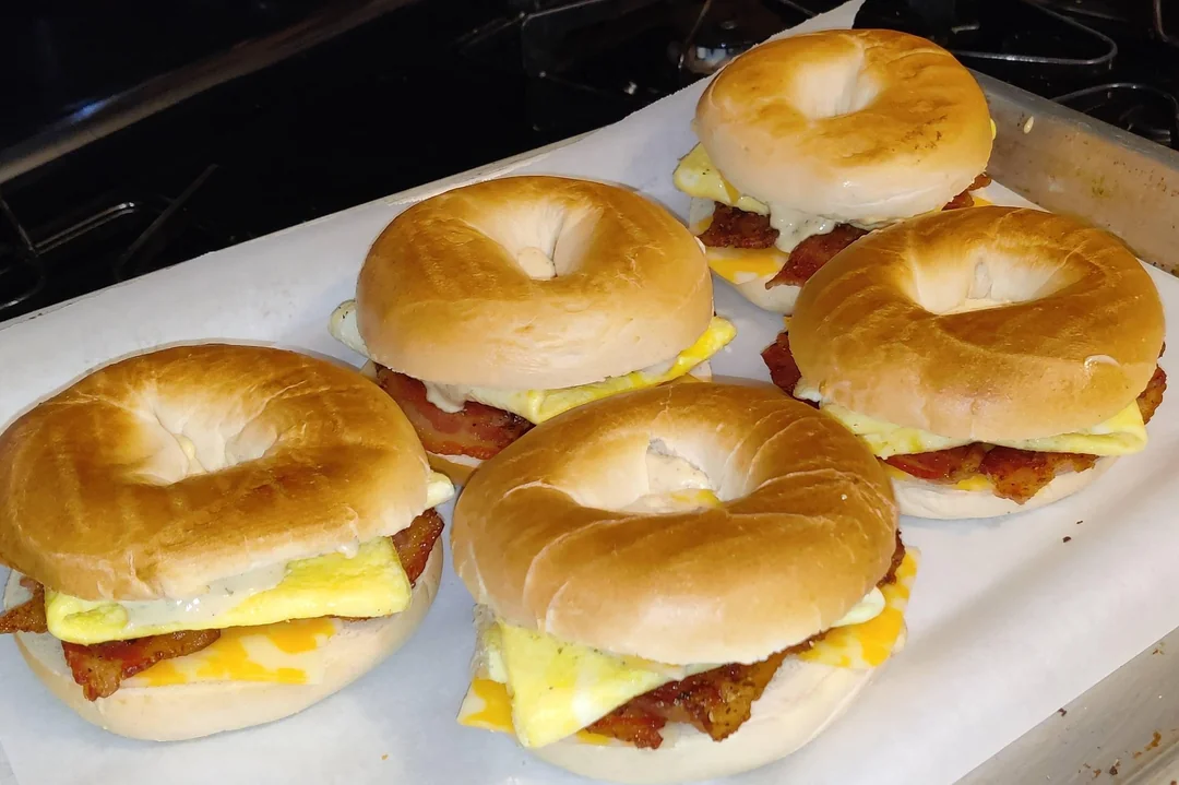 Five bagel breakfast sandwiches filled with egg, cheese, and bacon sit on a parchment-lined baking tray, ready to eat.