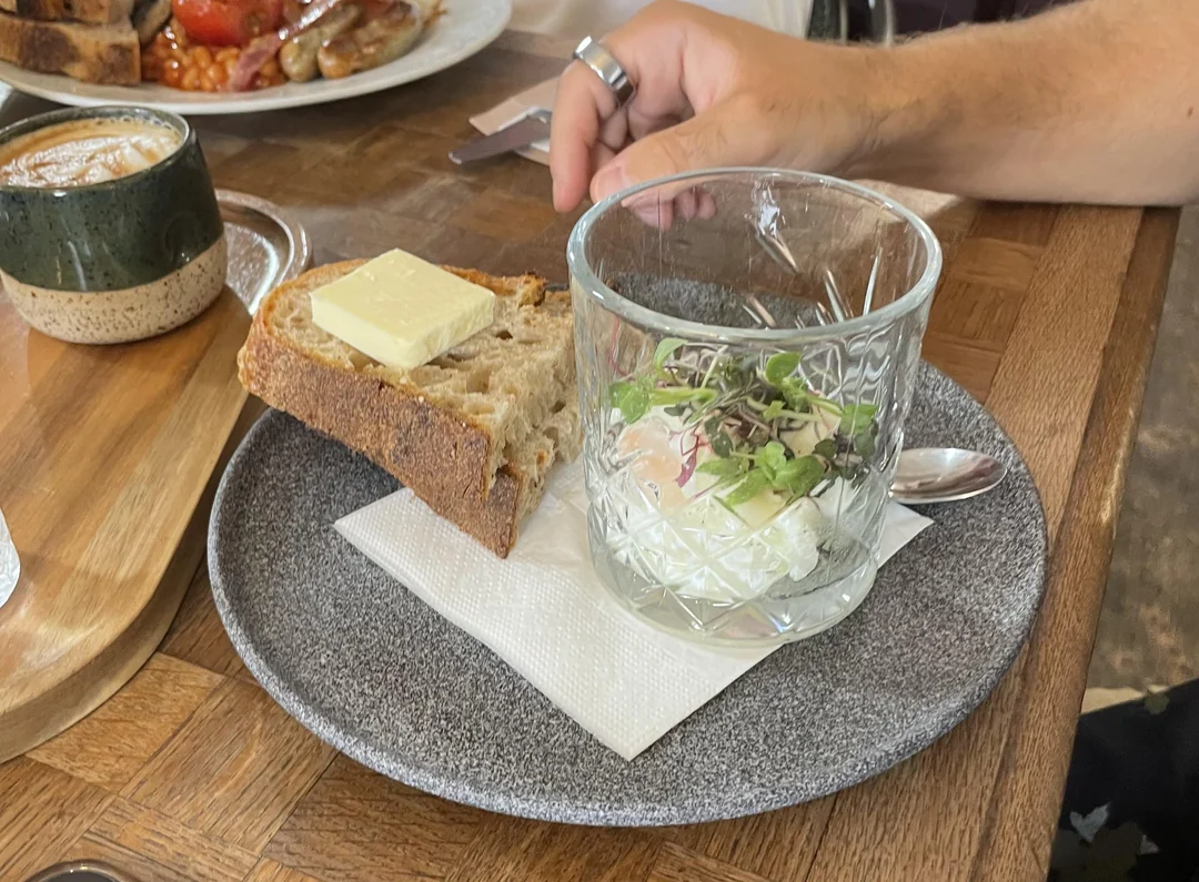 A plate with a slice of bread topped with butter, a glass containing a salad with microgreens, and a hand holding a fork, next to a cup of coffee on a wooden table.