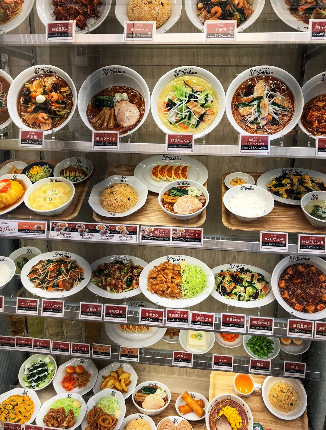Display case filled with realistic plastic models of various Japanese and Asian dishes, including ramen, rice bowls, stir-fried noodles, curry, salads, and side dishes, each labeled with name tags and prices.