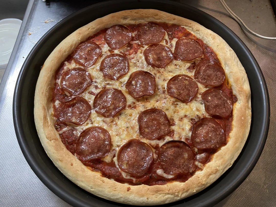 A freshly baked pepperoni pizza with melted cheese sits in a round black pan on a kitchen counter. The crust is golden brown and the pepperoni slices are evenly distributed.
