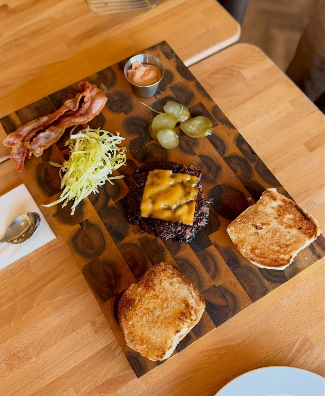 A deconstructed burger with a beef patty topped with melted cheese, toasted burger buns, shredded lettuce, bacon strips, pickles, and a small cup of sauce, all arranged on a wooden board.