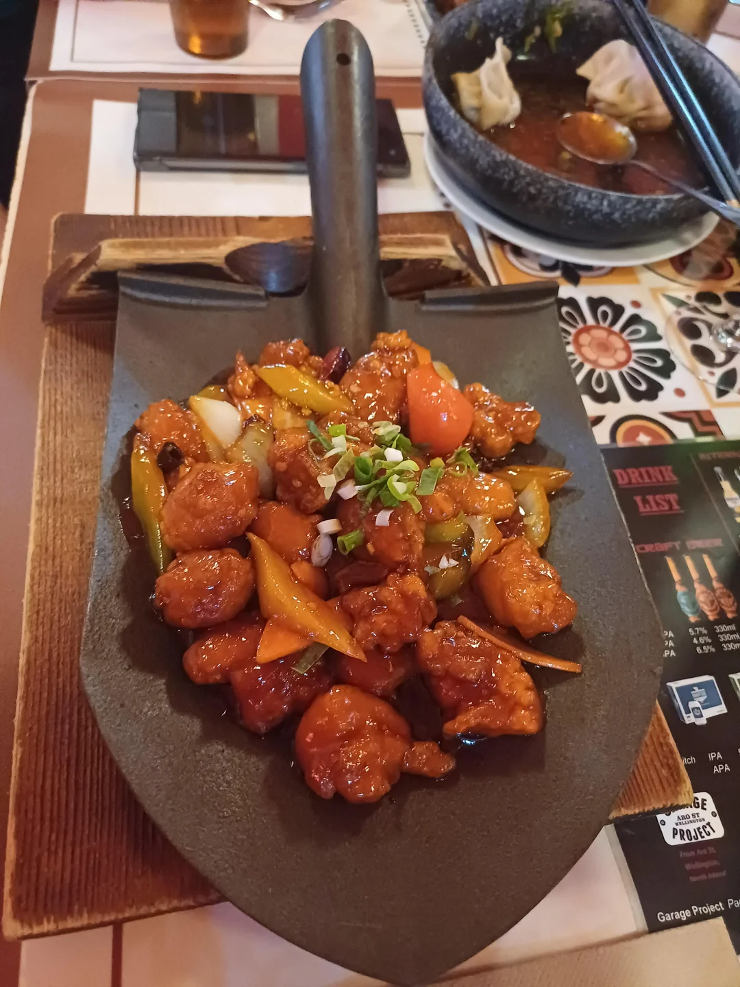 A dish of glazed chicken with vegetables served on a black shovel, garnished with green onions. A dumpling dish and drinks are in the background on a decorated table.