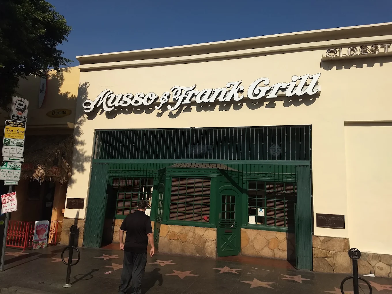 A person walks past the front of Musso & Frank Grill, a historic restaurant in Hollywood, with the Walk of Fame stars visible on the sidewalk.