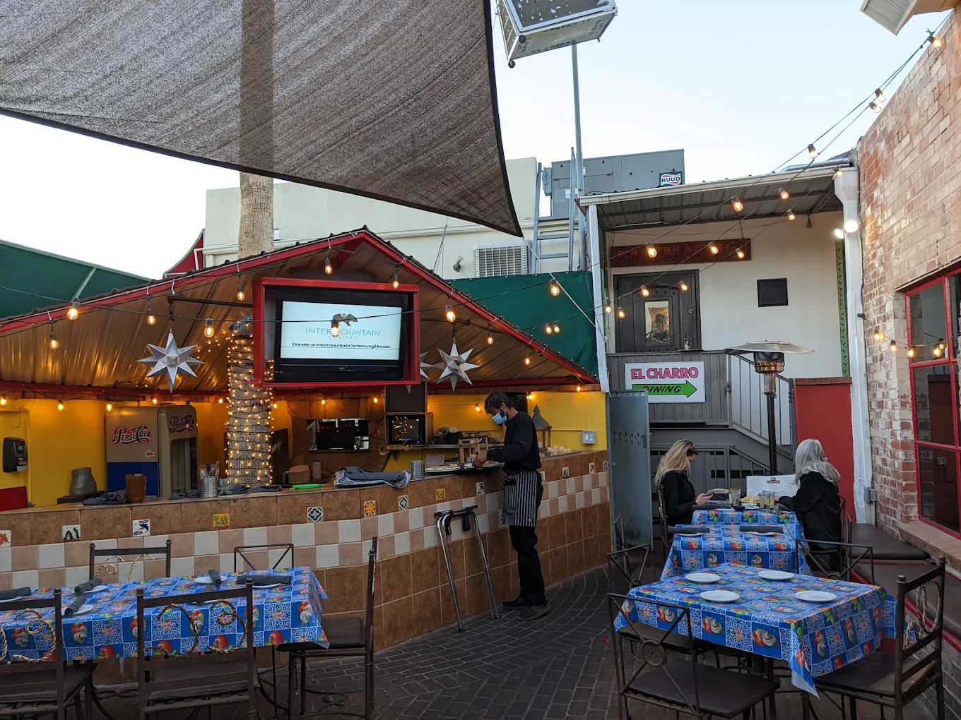 Outdoor restaurant patio with colorful tablecloths, string lights, and a counter decorated with holiday lights. A person stands at the counter, and two people sit at a table near a sign reading “El Charro.”