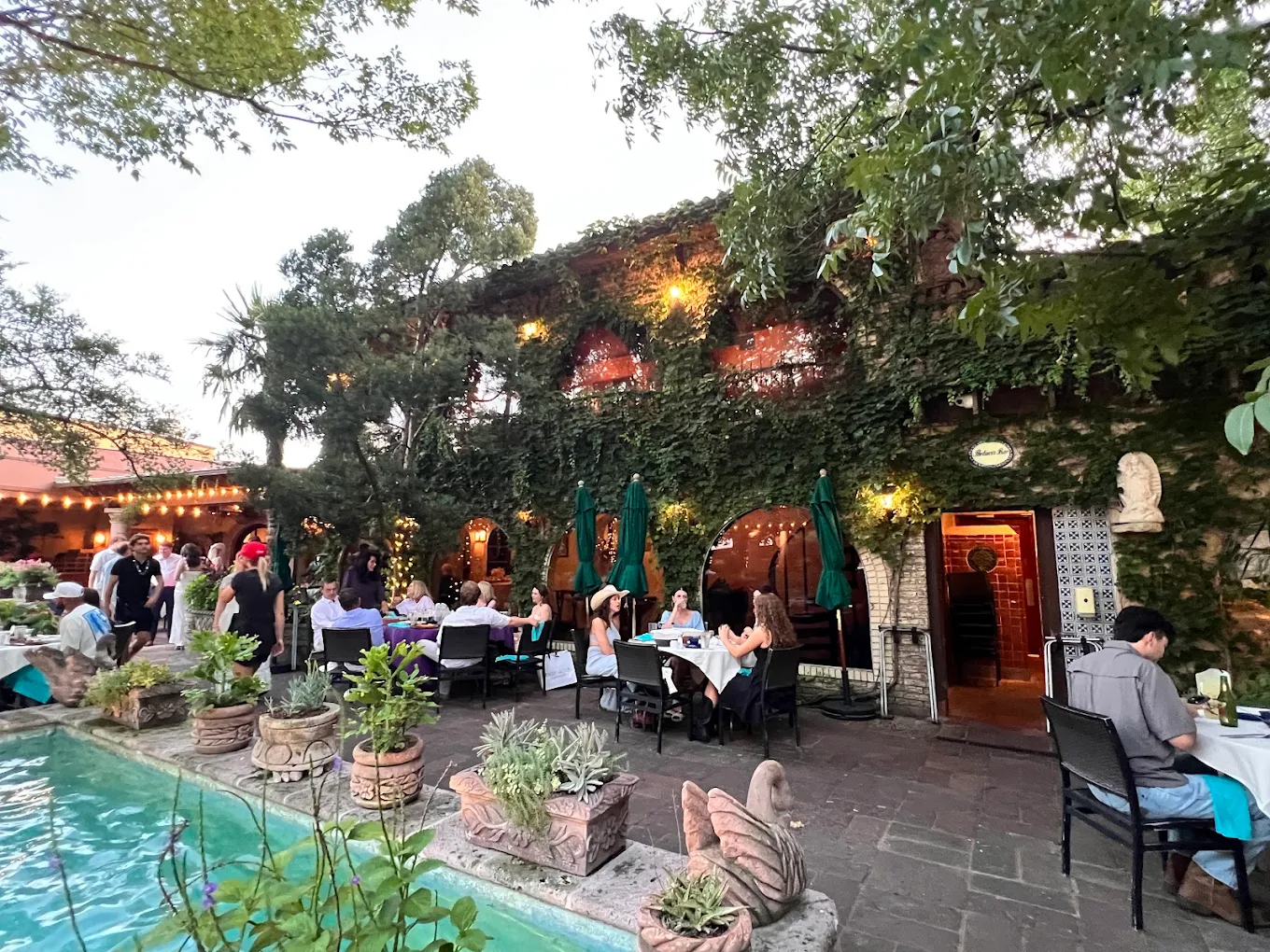Outdoor restaurant scene with diners seated at tables near a rectangular pool, surrounded by lush greenery and string lights. The building is covered in ivy, and trees create a cozy atmosphere.