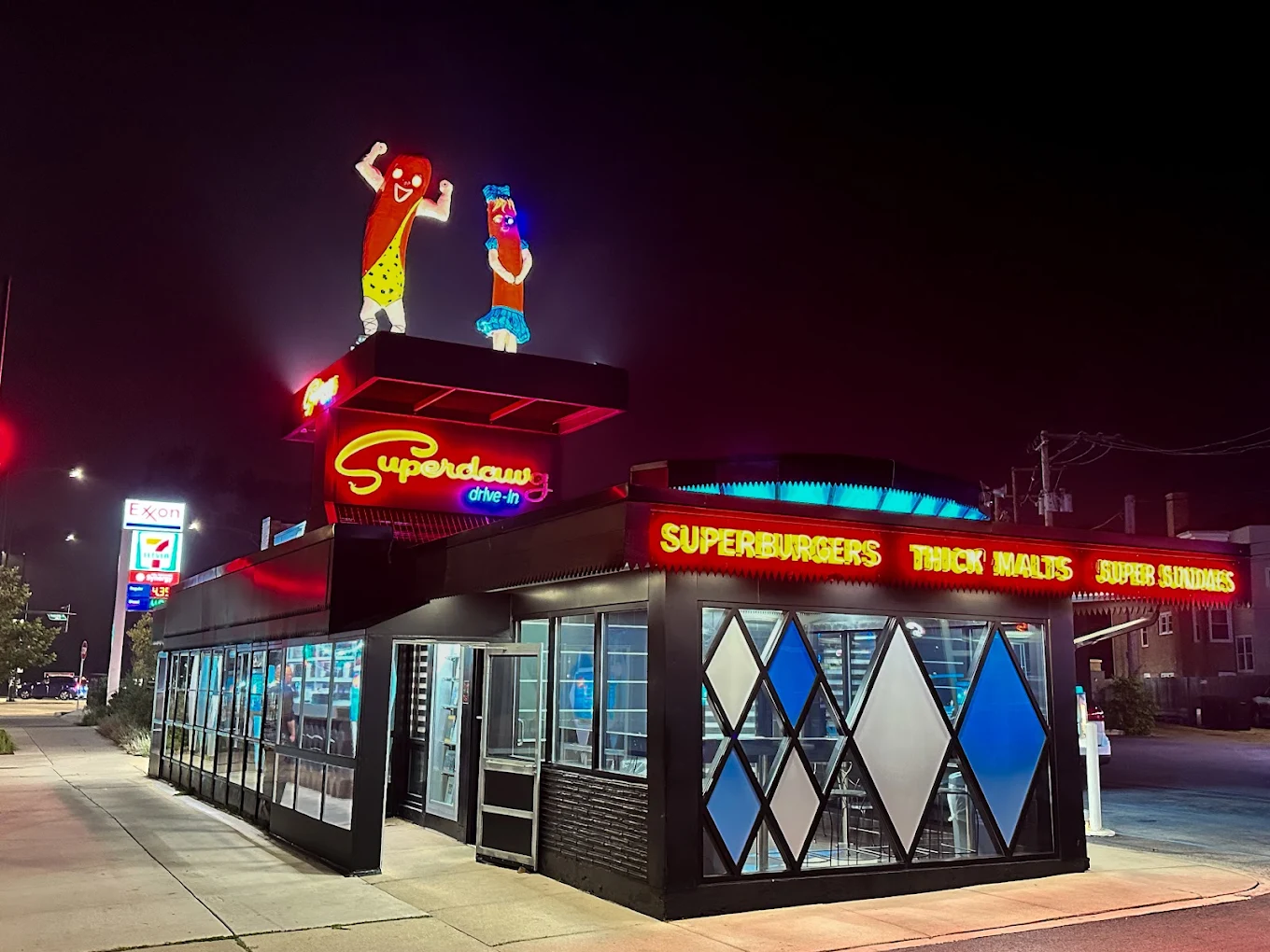 A retro diner at night with neon signs reading “Superdawg” and “SUPERBURGERS THICK MALTS SUPER SUNDAES.” Two hot dog mascots stand atop the roof. The diner has diamond-shaped windows and bright exterior lighting.