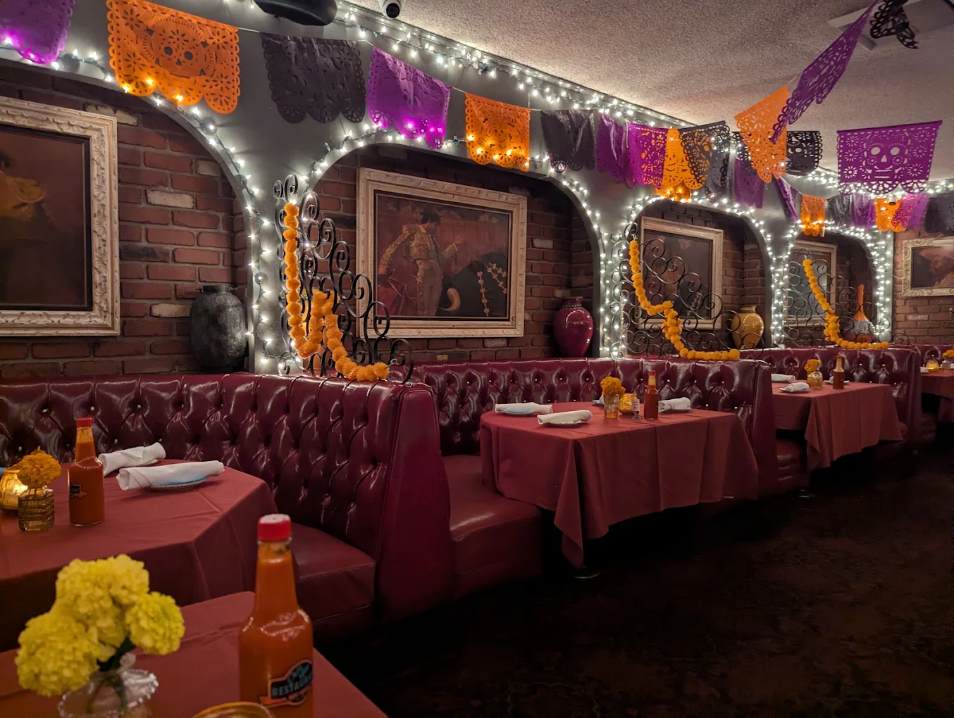 A cozy restaurant with red booths, tables set with yellow flowers, string lights, and colorful papel picado banners hanging above, with paintings on a brick wall.