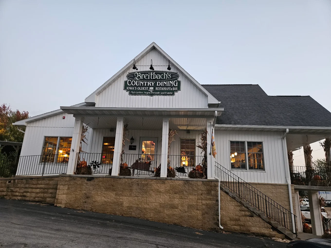 A white, two-story country restaurant named Breitbach’s Country Dining, with a porch, lights glowing inside, and autumn decorations on the steps and railings.