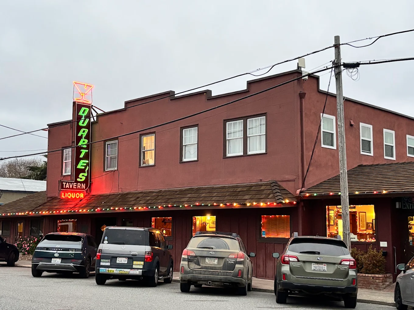 A brown tavern with neon signs reading “Quarters Tavern Liquor,” four cars parked in front, and string lights along the roof, under a cloudy sky.