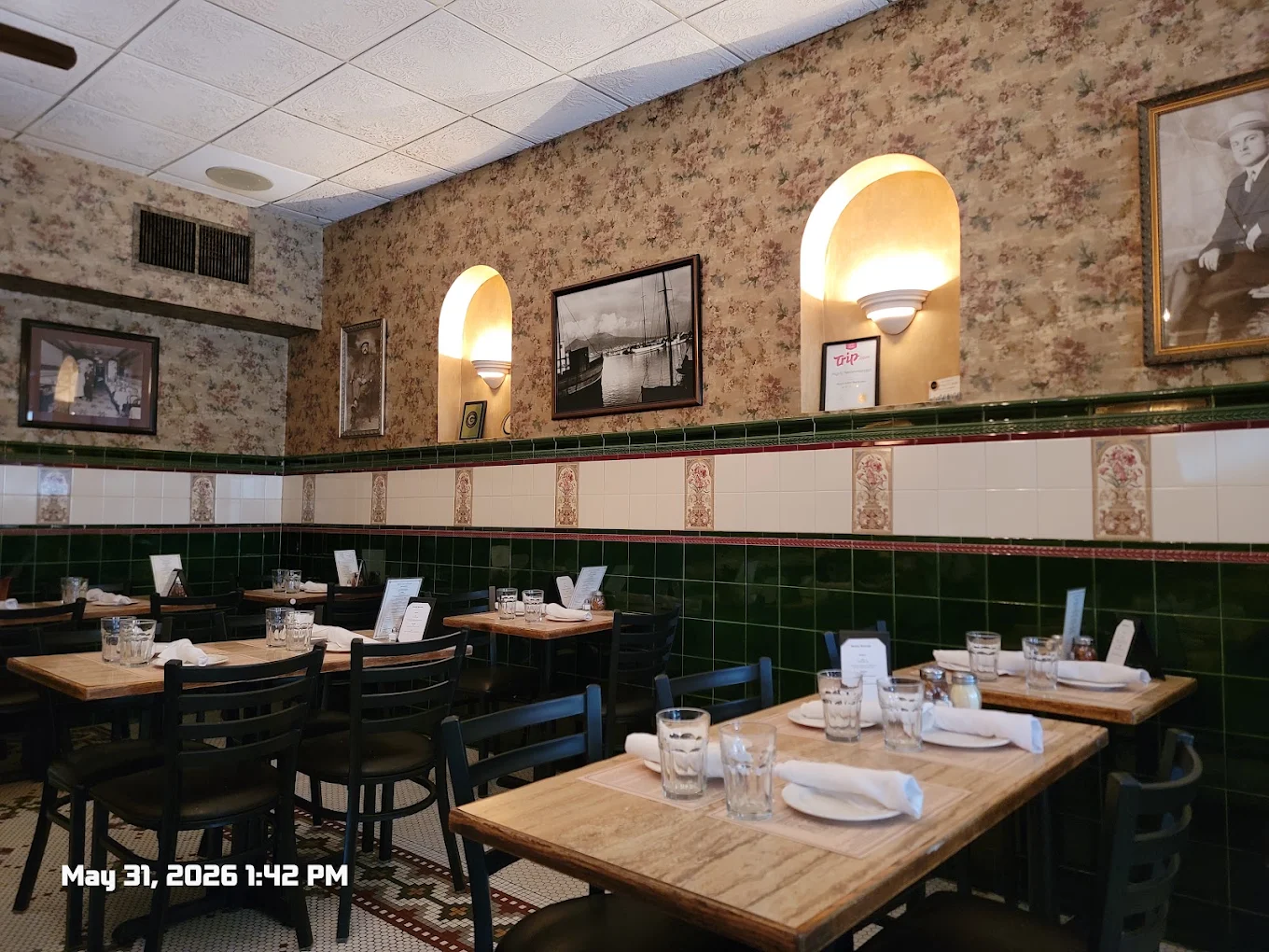 Cozy restaurant with floral wallpaper, framed black-and-white photos, empty wooden tables set with glasses and napkins, green and white tiled walls, and warm wall sconces. Timestamp: May 31, 2026, 1:42 PM.