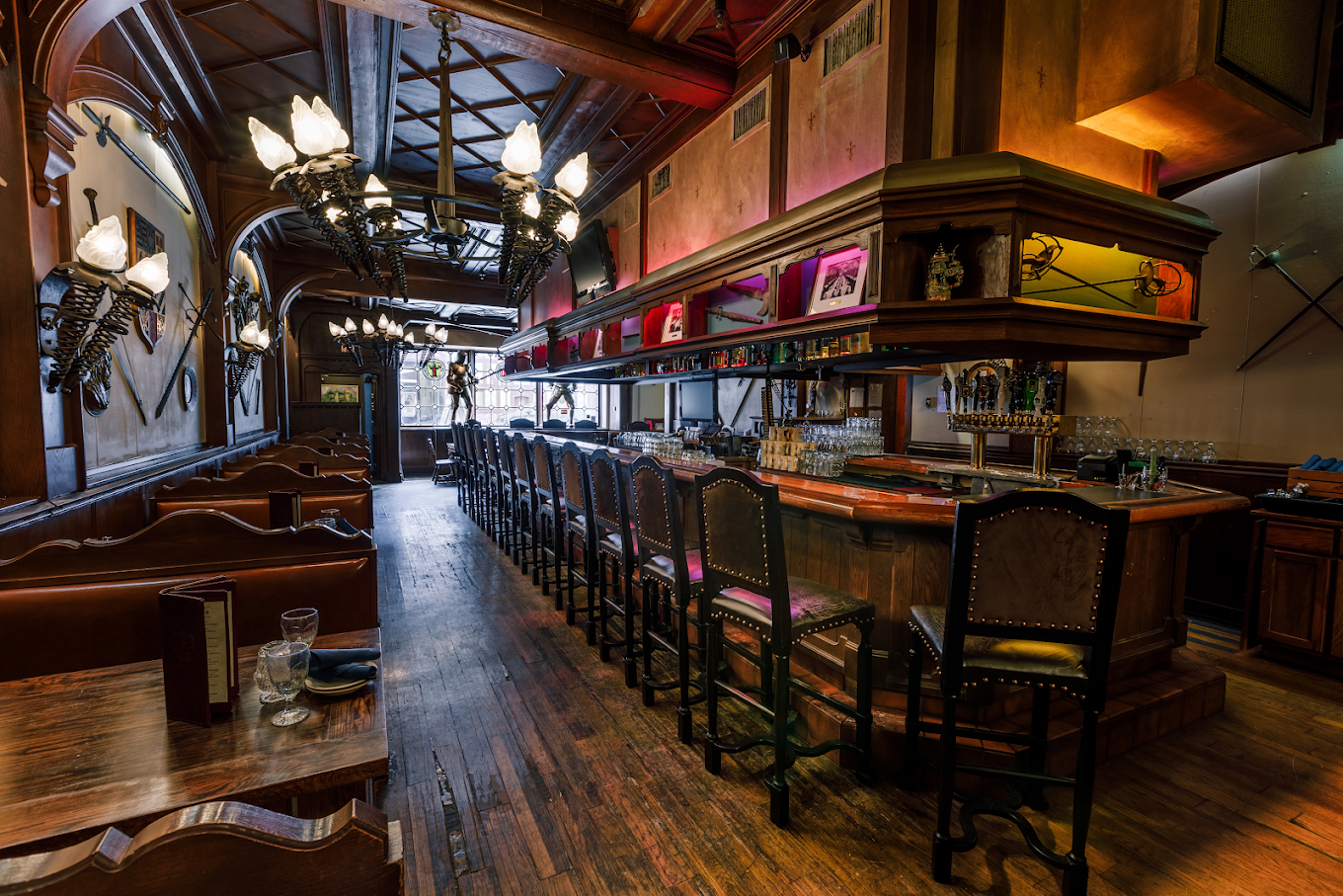 A rustic bar interior with wooden booths, a long bar counter lined with high chairs, shelves of bottles, ornate lighting, and decorative wall accents. Empty glasses sit on tables, and natural light shines in from windows.