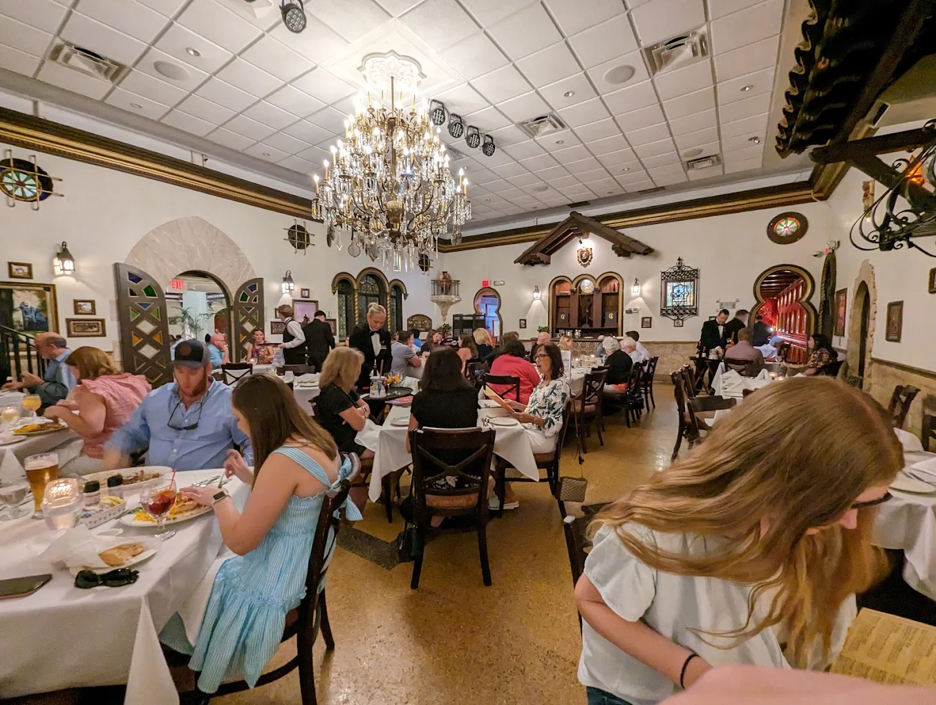 A busy restaurant dining room with people sitting at tables, eating and talking. A large chandelier hangs from the ceiling, and the decor is ornate with paintings and arches on the walls.