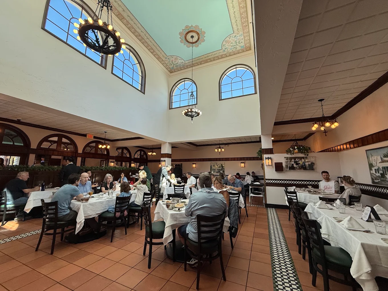 A spacious restaurant with high ceilings, large arched windows, and several groups of people dining at tables covered with white cloths. Warm lighting and classic decor create an inviting atmosphere.