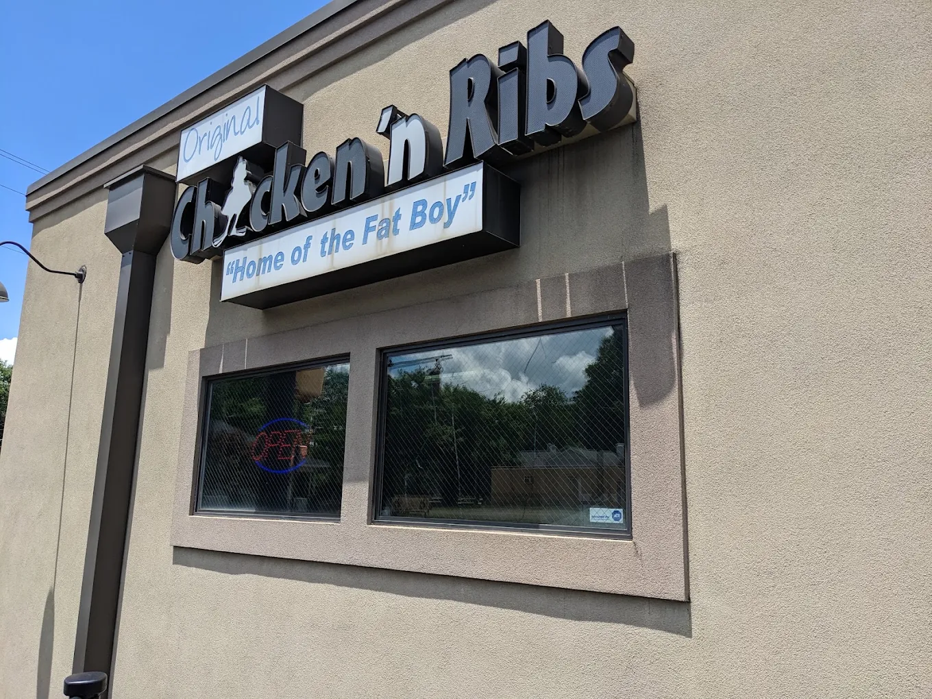 A beige building with signs reading “Original Chicken ’n Ribs” and “Home of the Fat Boy.” There are two windows, one displaying a lit “OPEN” sign. Trees and sky are reflected in the glass.