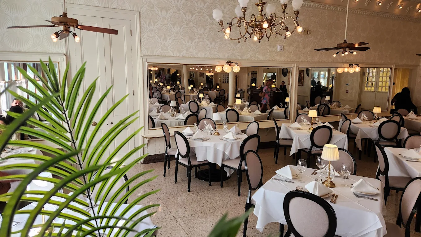 Elegant restaurant with round tables covered in white cloths, set for dining. Large mirrors line the walls, chandeliers hang from the ceiling, and a leafy plant is in the foreground. Several people are visible in the background.