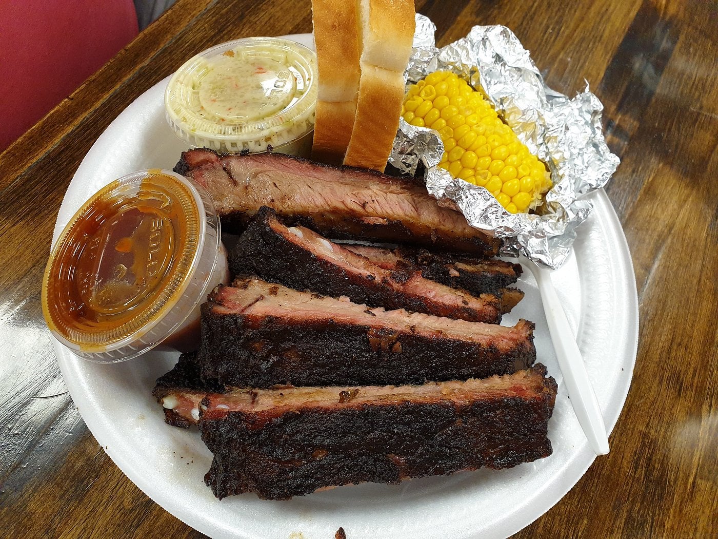 A plate of barbecue ribs, corn on the cob in foil, coleslaw, barbecue sauce, and two slices of white bread on a white foam plate atop a wooden table.