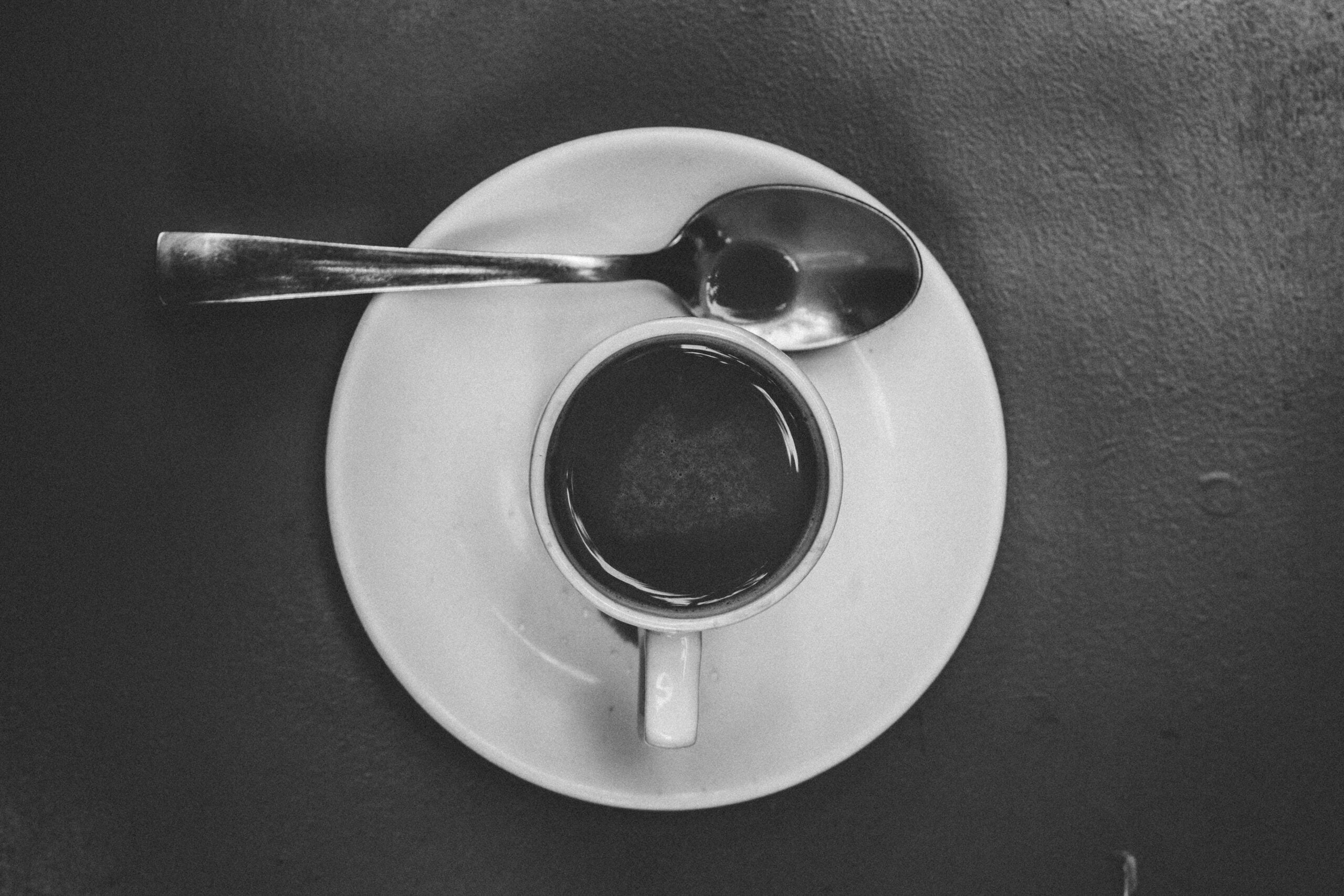 A small cup of coffee on a white saucer with a spoon resting across the top, photographed from above in black and white.