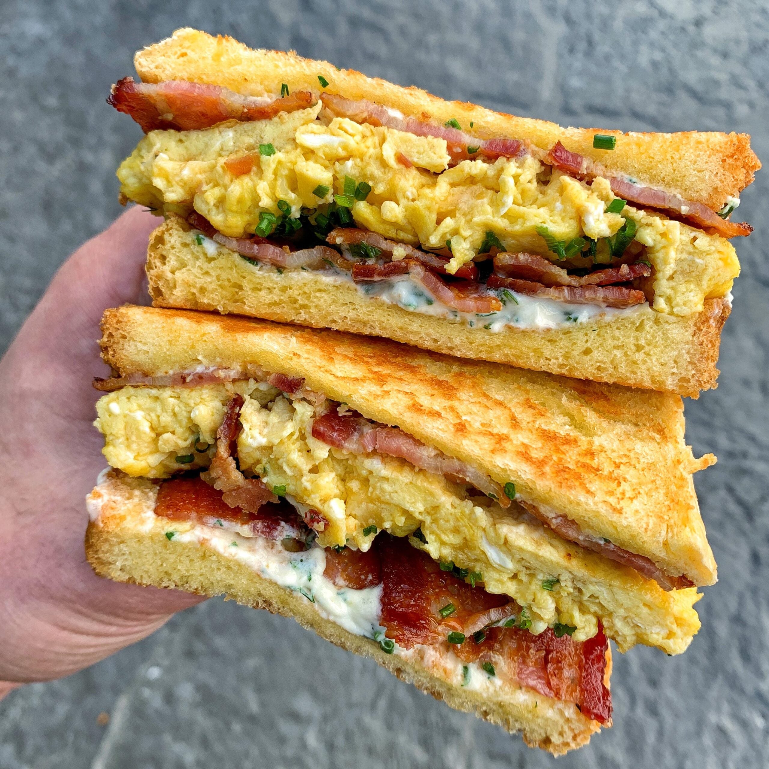 A hand holds two stacked breakfast sandwiches filled with scrambled eggs, crispy bacon, fresh chives, and a creamy sauce, all between toasted slices of bread.