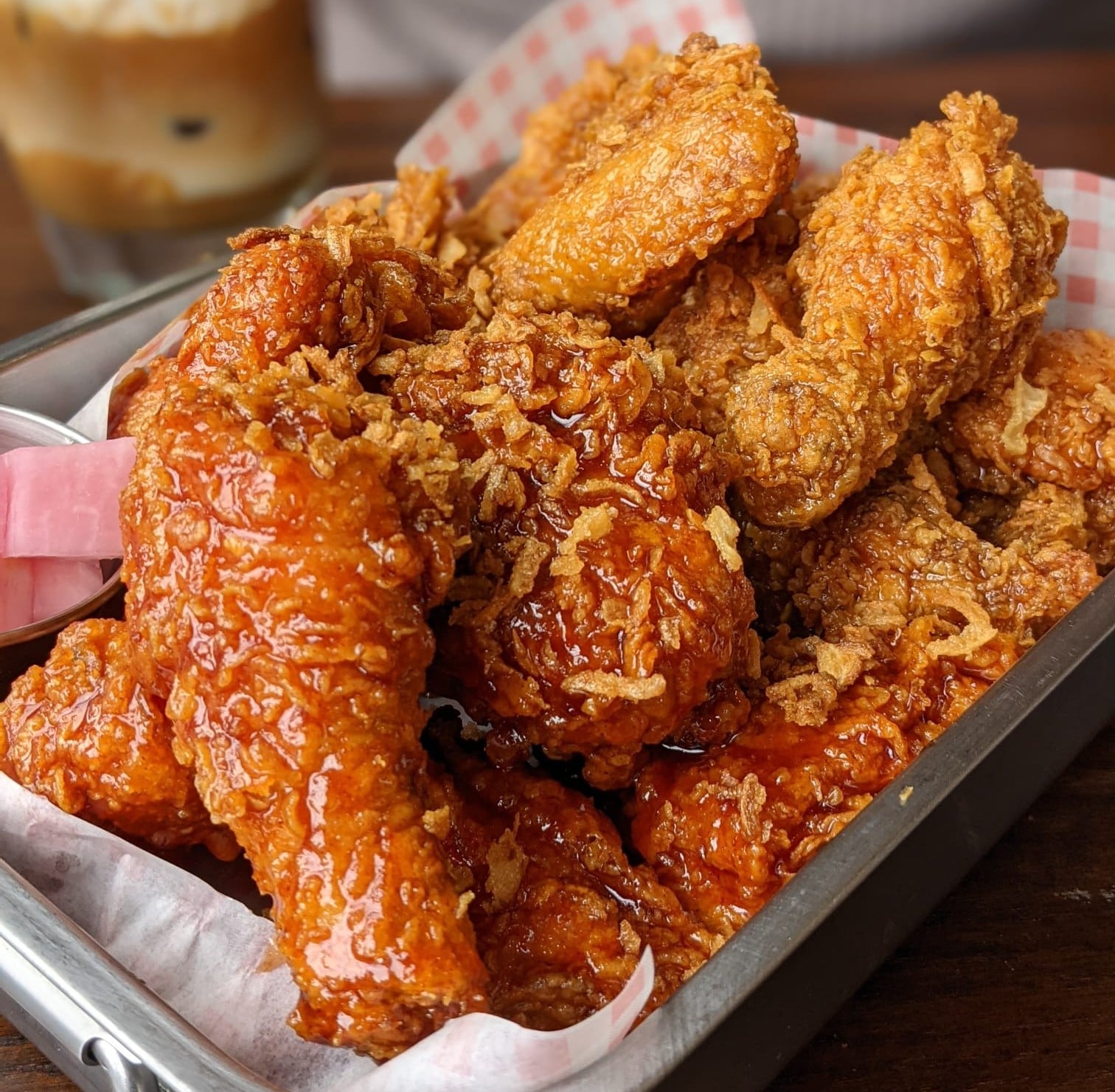 A metal tray filled with crispy fried chicken wings, some glazed with a reddish sauce and others plain, sits on a table. A drink with ice is blurred in the background.