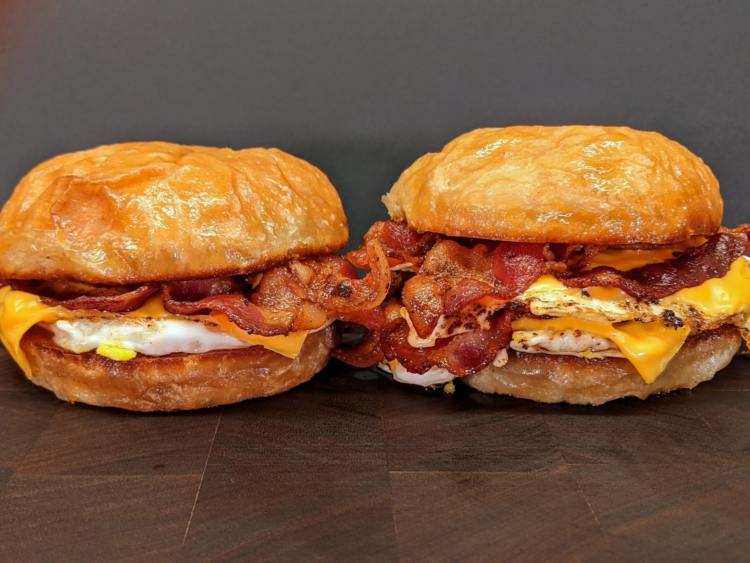 Two croissant sandwiches filled with crispy bacon, egg, and melted cheese, placed side by side on a dark wooden surface with a plain dark background.